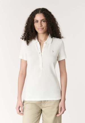 Donna con capelli scuri e ricci che indossa una polo bianca a maniche corte e pantaloni beige, in piedi davanti a uno sfondo chiaro e uniforme.