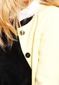 Capelli biondi sopra una maglia nera con un collo bianco, parzialmente coperti da una giacca giallo pallido con due bottoni neri visibili.