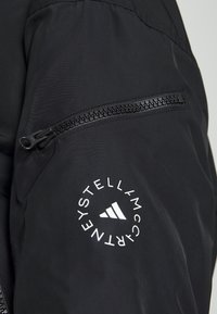 Black jacket featuring a zippered pocket on the sleeve, with a white circular logo and three-stripe graphic detailing. Smooth fabric texture.
