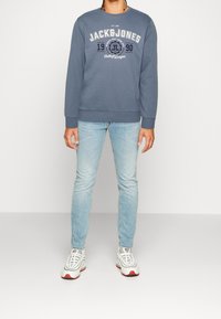 Felpa blu con "JACK & JONES" stampato in bianco e blu marino, abbinata a jeans azzurri e sneakers bianche con dettagli rossi.