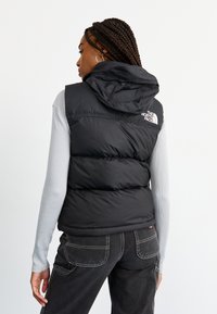 Vestă puffer neagră cu glugă, având un logo de marcă pe umăr. Asortată cu un top cu mâneci lungi de culoare gri deschis și blugi negri.