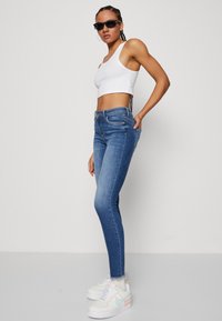 Žena v čiernych slnečných okuliaroch, bielom crop tope, modrých skinny džínsoch a bielych teniskách stojí proti jednému svetlému pozadiu.