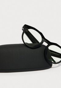 Mustat suorakulmaiset aurinkolasit mustassa pehmeässä kotelossa. Aurinkolasit ovat varustettu kirkkailla linsseillä ja kulmikkaalla, geometrisella kehysmuotoilulla.