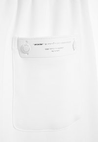 Prenda blanca con un bolsillo que tiene una etiqueta plateada. La etiqueta contiene texto en relieve y detalles metálicos. El material parece liso y texturizado.
