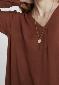 Bruine V-hals top van textuurstof, losse pasvorm en lange mouwen. Beschikt over een kleine insnoering en knoopdetails bij de manchetten. Een gouden ronde hanger ketting.