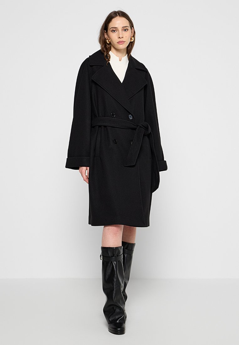 Czarny płaszcz z wełny w stylu trencza, z podwójnym zapięciem, szerokimi klapami, paskiem w talii i długimi rękawami, połączony z czarnymi kozakami do kolan.
