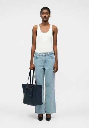 Femme portant un débardeur blanc, un jean bleu clair à jambes larges, des talons noirs, tenant un grand sac cabas noir, debout devant un fond blanc uni.