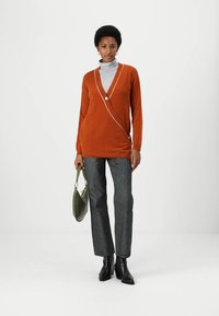 Langärmlige orange Strickjacke mit weißer Paspelierung und einem knöpfbaren Verschluss. Getragen über einem grauen Rollkragenpullover und kombiniert mit dunkelgrauen Hosen und schwarzen Stiefeletten.