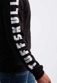 Felpa nera con cappuccio con scritta audace e bianca "TUFF SKULL" lungo la manica sinistra. Il tessuto appare liscio, con una vestibilità regolare e senza decorazioni.