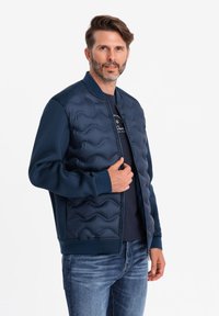 Veste de bombardier marine avec un devant matelassé ; manches et col lisses ; poignets côtelés ; fermeture éclair ; portée par-dessus un t-shirt noir et un jean.
