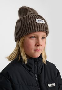 Bruine gebreide muts met een strakke pasvorm en geribbelde textuur, met een wit label met "hummel sportswear" aan de voorkant. Zwarte gewatteerde jas zichtbaar.