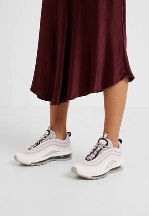 Nogi w białych i szarych sneakersach Nike Air Max 97 z czarnymi sznurówkami i taśmami na pięcie, zestawione z lejącą się bordową spódnicą.