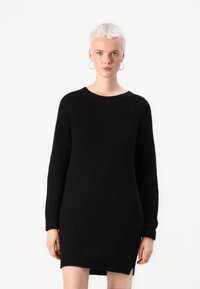 Robe en tricot noir avec texture côtelée, col rond et manches longues. Fentes latérales au niveau de l'ourlet. Le tissu semble épais et chaud.