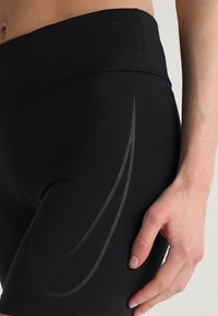 Shorts de sport noirs avec une taille haute, arborant un logo swoosh gris subtil sur le côté. Fabriqués en tissu extensible et lisse.
