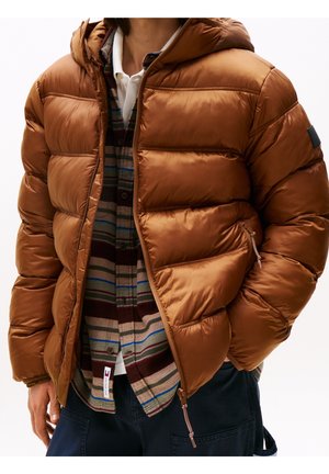 Homme portant une doudoune marron par-dessus une chemise rayée et un sous-vêtement blanc avec un pantalon sombre, debout avec une main dans la poche.