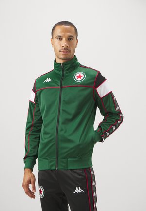 Veste de sport verte avec col haut, fermeture éclair à l'avant, accents rouges et blancs, et un écusson de logo. Associée à un pantalon noir avec des rayures sur les côtés.