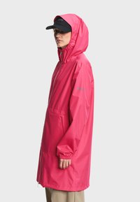 Femme portant un imperméable à capuche rose vif avec une fermeture éclair à l'avant et des poches, associée à une casquette de baseball noire, sur un fond uni.