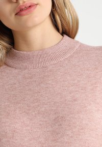 Světle růžový svetr s pleteným kulatým výstřihem a měkkou, texturovanou tkaninou. Detailní pohled zvýrazňuje jemné variace v barvě a textuře.