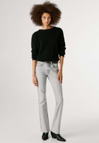 Μαύρο πλεκτό πουλόβερ με φαρδιά μανίκια, συνδυασμένο με ανοιχτό γκρι τζιν με καμπάνα και μαύρα μποτάκια με τακούνι, σε απλό φόντο.