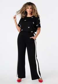 Femme aux cheveux bouclés portant une chemise noire avec des nœuds blancs, un pantalon noir avec des bandes blanches sur les côtés, et des chaussures rouges pointues, posant avec une main dans la poche.