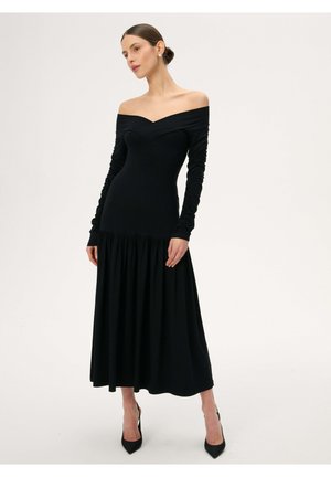 Kobieta ubrana w czarną, długą sukienkę midi z odkrytymi ramionami i marszczonymi rękawami, oraz czarne szpilki z ostrym noskiem, stojąca na jednolitym tle.