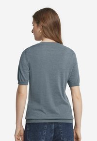 Kvinna med långt brunt hår som bär guldörhängen, en grå kortärmad tröja och mörkblå jeans, sedd bakifrån.