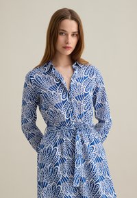 Abito blu a maniche lunghe con un motivo astratto bianco, caratterizzato da un collo a camicia, chiusura con bottoni e una cintura abbinata in vita.