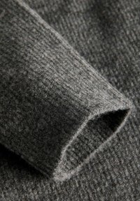 Tessuto grigio con un motivo tessuto e testurizzato. Il primo piano mostra un bordo della manica curvo, mettendo in risalto le morbide e pelose fibre.