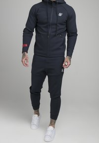 Mörkblå, zip-up huvtröja med en huva och sidofickor, kombinerad med matchande joggers. Har en logotyp och röda detaljer på ärmen. Vita sneakers.