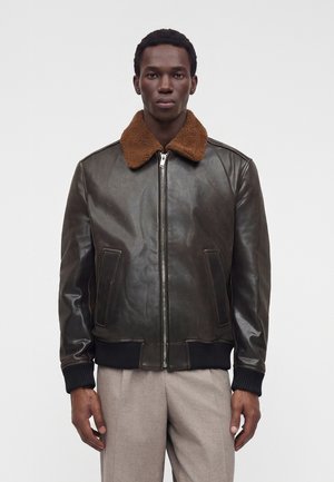 Mand iført en mørkebrun læderjakke med brun shearling-krave og lyse beige bukser, stående mod en ensfarvet baggrund.
