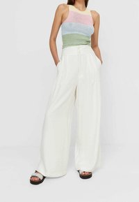 Vrouw in een mouwloos gestreept gehaakt topje en hoge, brede witte pantalons met haar handen in de zakken, staand tegen een witte achtergrond.