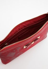 Červená kabelka typu clutch s texturovaným povrchem, zlatým zipem, vnitřními kapsami a dekorativní mašlí na přední straně.