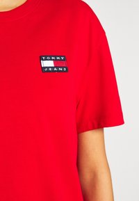 Czerwony t-shirt z krótkim rękawem z małą naszywką logo Tommy Jeans na lewej piersi, widoczna część górnej sylwetki osoby.