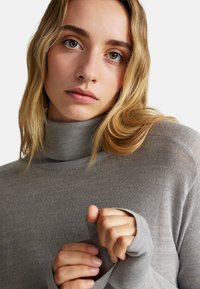 Maglione grigio a collo alto realizzato in un tessuto morbido e texturizzato con polsini aderenti. Il materiale appare leggero e confortevole, con una sottile lavorazione a coste.