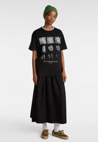 Camiseta negra con estampado gráfico blanco que presenta varios rostros, llevada con una larga falda negra, calcetines blancos y zapatillas marrón con manchas.