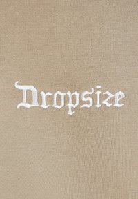 Beiger Stoff mit weißem gesticktem Text "Dropsize" und dekorativen Verzierungen auf einem glatten, gleichmäßigen Hintergrund.