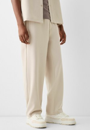 Homme portant un pantalon beige à jambes larges, des baskets blanches, et une chemise beige boutonnée par-dessus un t-shirt taupe, sur un fond uni.