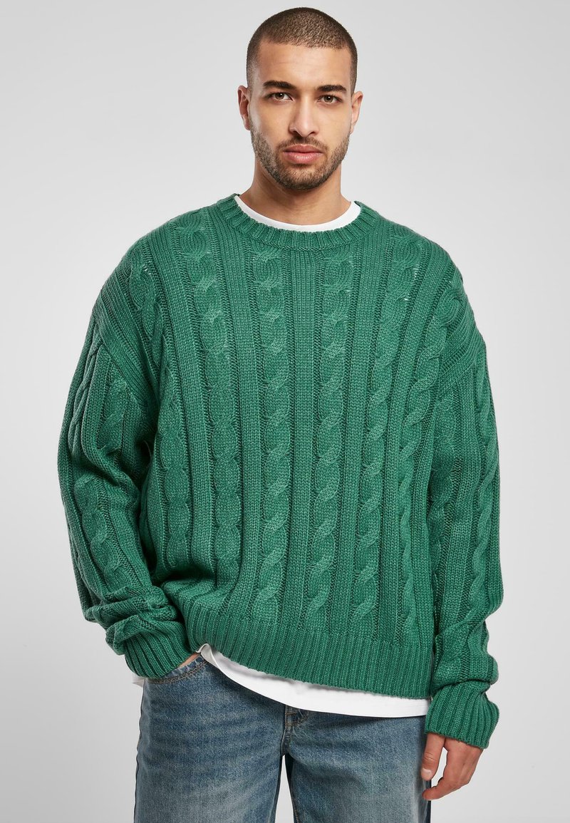 Green cable-knit sweater with a loose fit, ribbed cuffs, and hem. Worn over a white t-shirt, featuring textured patterns throughout.