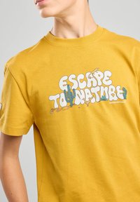 Camiseta de algodón amarilla con un diseño gráfico que presenta el texto "ESCAPA A LA NATURALEZA" e ilustraciones de cactus y animales en blanco y verde.