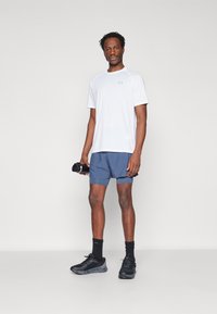 Camiseta atlética blanca, pantalones cortos azules, zapatillas negras, sosteniendo una botella de agua. Presenta mangas cortas, tela ligera y un diseño ajustado.