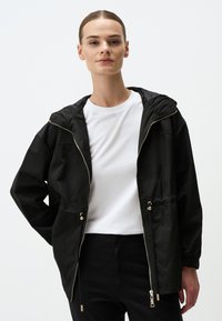 Zwarte hoodiejas met een ritssluiting aan de voorkant, elastische manchetten en een trekkoord in de taille. De stof lijkt glad en lichtgewicht. Eenvoudig ontwerp.