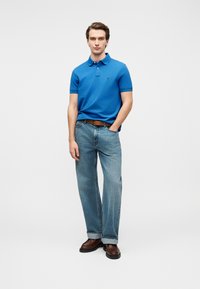 Polo bleu, jean en denim, ceinture marron et chaussures marron. Le polo comporte un col et des manches courtes, le jean a une coupe décontractée et une teinte claire.