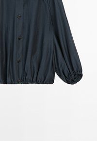 Mörk marinblå blus med långa ärmar, knäppning fram, rynkade manschetter och lätt rynkad fåll på vit bakgrund.