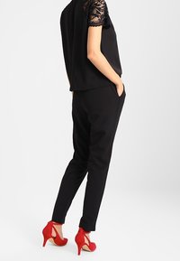 Conjunto negro que incluye una blusa con mangas de encaje, pantalones negros ajustados y zapatos de tacón rojos con un diseño recortado en la parte trasera.