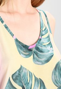 Närbild av en person som bär en gul linne med stora gröna tropiska bladtryck och en Adidas-logotyp nära halslinningen.