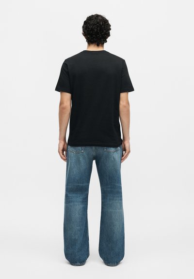 Άνδρας με σγουρά σκούρα μαλλιά που φοράει απλό μαύρο t-shirt και φαρδιά μπλε τζιν, στέκεται με την πλάτη σε λευκό φόντο.