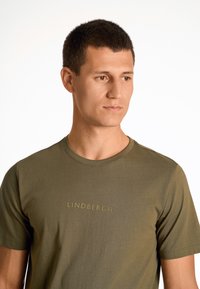 Camiseta de algodón verde oliva con cuello redondo y mangas cortas, con "LINDBERGH" impreso en amarillo claro en el pecho.