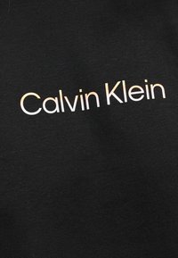 Μαύρο ύφασμα με λευκό και ανοιχτό πορτοκαλί κείμενο σε γραμμάρια βαθμίδας που γράφει «Calvin Klein» σε απλό γραμματοσειρά sans-serif.