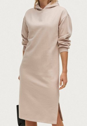 Kvinde iført en beige, langærmet hættetrøje-kjole med sideslids, stående mod en ensartet lys baggrund.