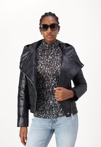 Zwarte gewatteerde pufferjack met een brede kraag, ritssluiting, gedragen over een gemusterde zwart-grijze coltrui, gecombineerd met lichtblauwe jeans.